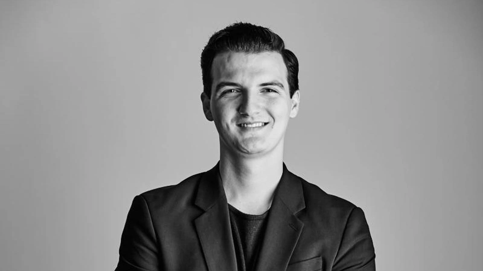 Black-and-white portrait of Premtim Gjonbalic smiling in a blazer against a plain background.